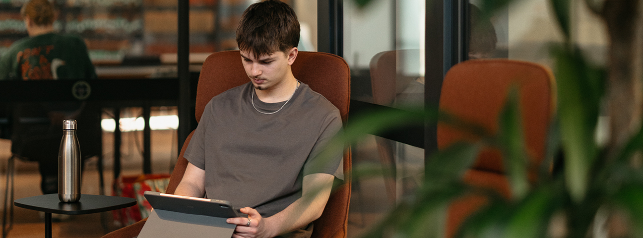 Male computer science student sitting inside a library in a chair working on his iPad. 
