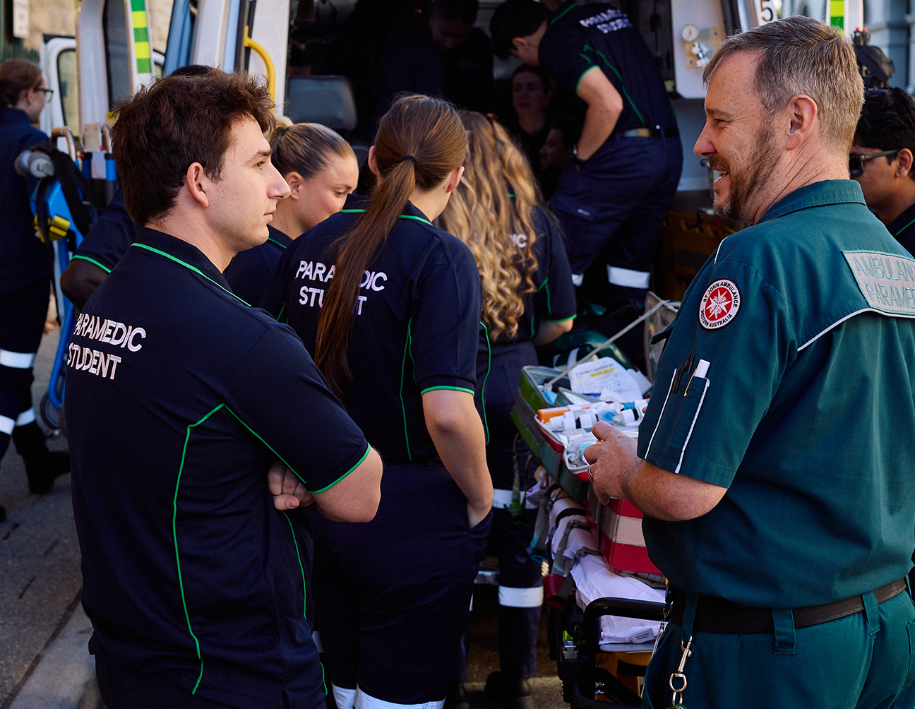 A group of UNDA paramedicine students listening and working with paramedics outside of an ambulance.