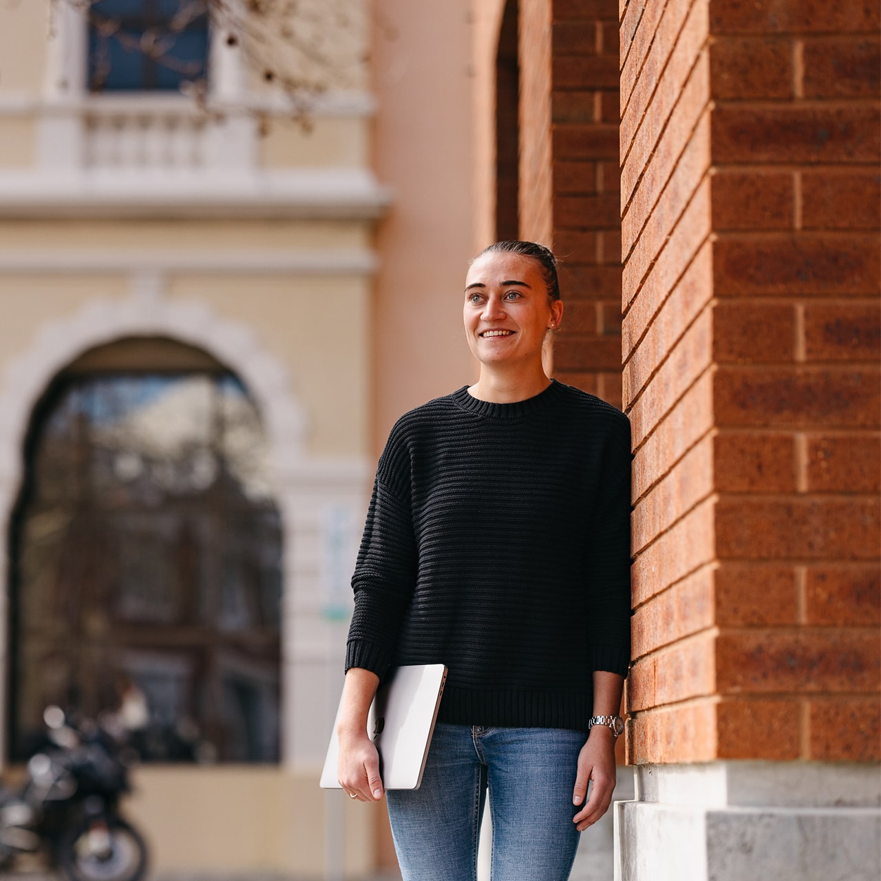 A female student wearing a black sweater leaning against a Fremantle campus building carrying her laptop 
