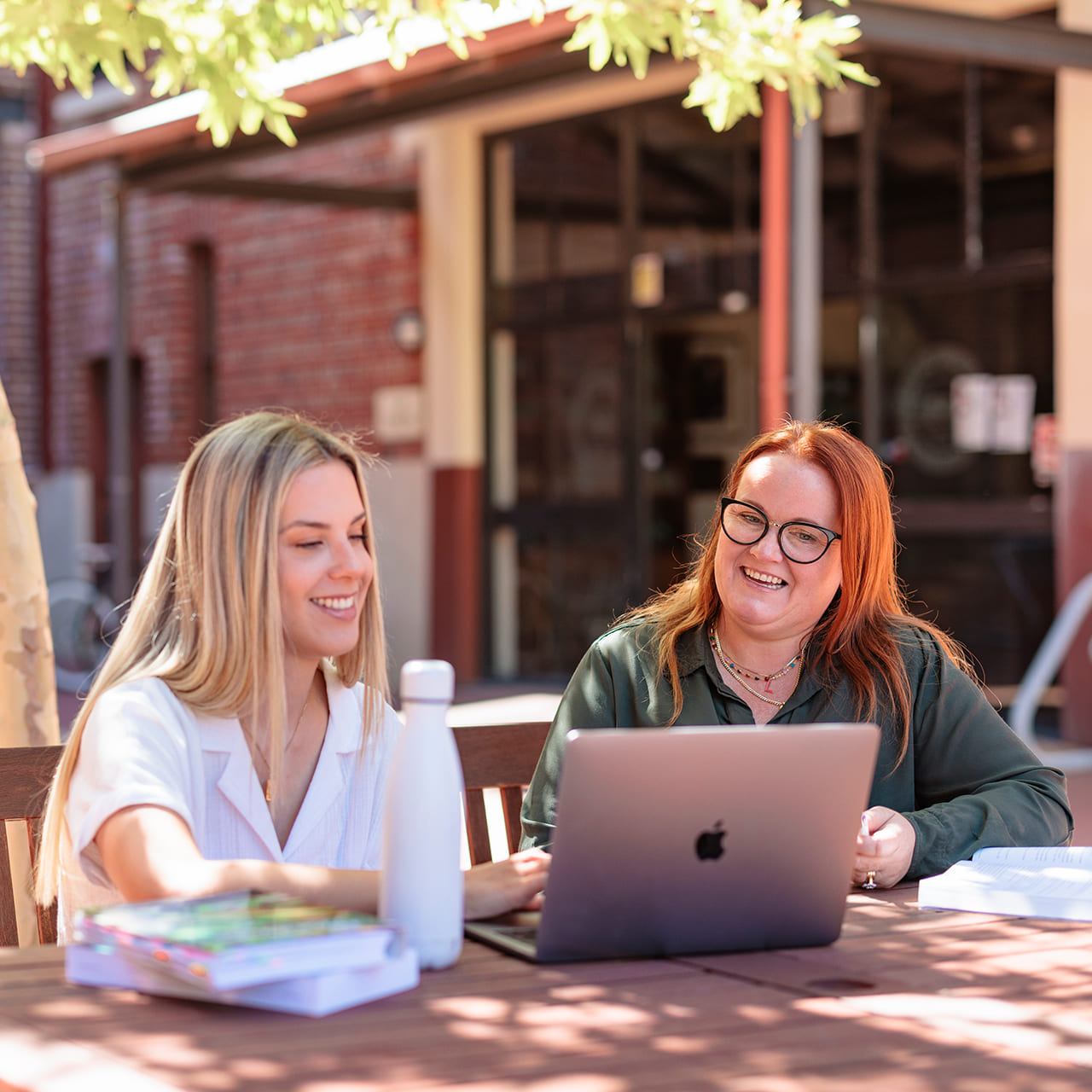 Two female students sitting down at a table in a campus courtyard looking at a laptop together 