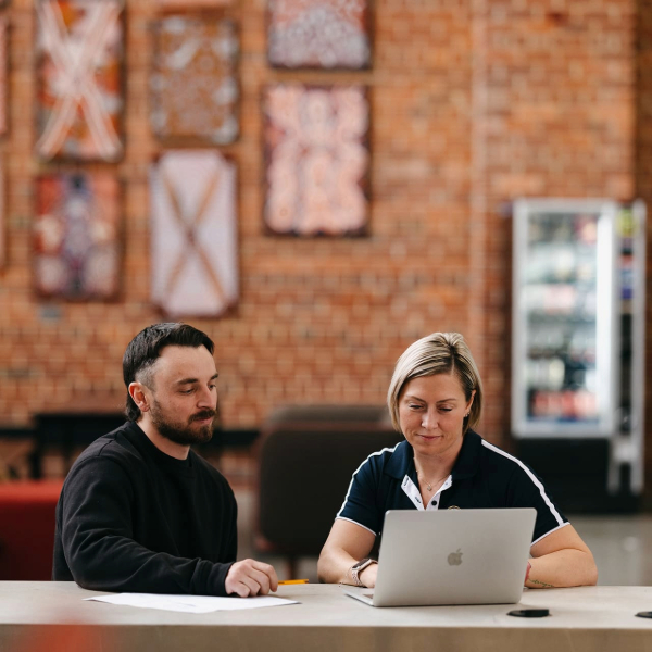 Two students sitting down at a table looking at a laptop together 