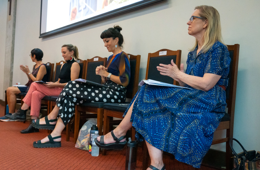 Four females sitting on wooden chairs in a row with papers on their laps and their hands in a prayer position