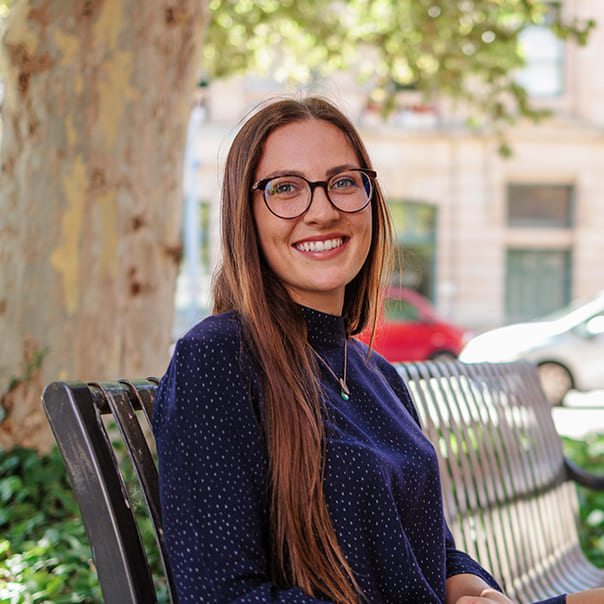 Female student with glasses in a dark blue turtleneck shirt sitting and smiling on a bench outside of campus 