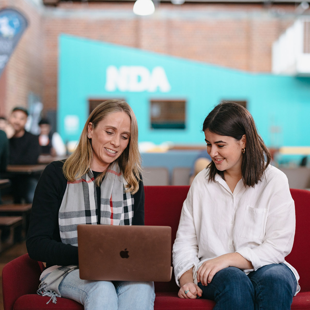 Two females postgraduate students using a laptop while sitting on a red couch 