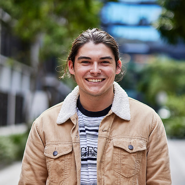 A smiling Notre Dame male student wearing a brown jacket standing outside in the Sydney campus 