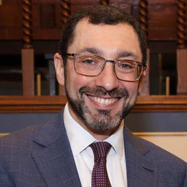 A man with glasses is facing the camera and smiling in front of a wooden bookcase
